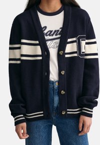 Granatowy kardigan w białe paski z logo, zapinany na guziki, z prążkowanymi mankietami i brzegiem, noszony z białą koszulką i niebieskimi dżinsami.