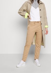 Beige Jogginghosen mit Kordelzug, kombiniert mit einem weißen Oberteil und einem grauen Reißverschluss-Sweatshirt unter einem hellbeigen Trenchcoat mit neon grünen Akzenten.