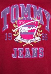 Tommy Jeans-Logo mit lila und weißen Buchstaben, einem roten und lila Wappen, goldenen Lorbeerblättern und dem Text "NYC USA" sowie "Since 1985" auf rotem Stoff.
