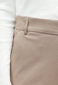 Beżowe spodnie o teksturowanej tkaninie, z gładkiego materiału, z szerokim pasem i subtelnymi bocznymi kieszeniami, w połączeniu z białą bluzką w prążki.