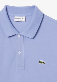 Lacoste-paita vaaleansinisestä, teksturoidusta kankaasta, jossa on kolmonen napitus ja pieni brodeerattu krokotiili-logo vasemmassa rinnassa.