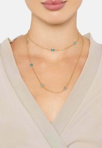 Gold-tone necklaces in layered design with blue rectangular accents. Chains are delicate with a smooth texture, worn over a beige garment.