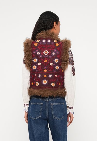 Prešívaná vesta s vínovou látkou, kvetinovou výšivkou a hnedým umelým kožúškom. Kombinovaná s bielou košeľou a modrými džínsami, zobrazená zozadu.
