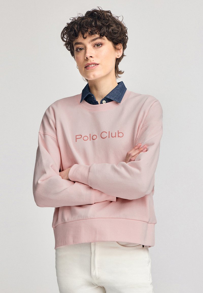 Felpa rosa con scollatura rotonda e polsini a coste. Presenta la scritta "Polo Club" in una tonalità leggermente più scura. Indossata sopra una camicia di jeans.