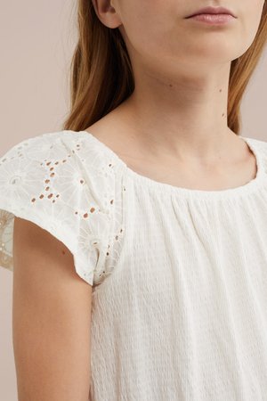 Mladić s ravnom smeđom kosom nosi bijelu majicu od teksturiranog tkanja s čipkastim kratkim rukavima s cvjetnim izrezima.