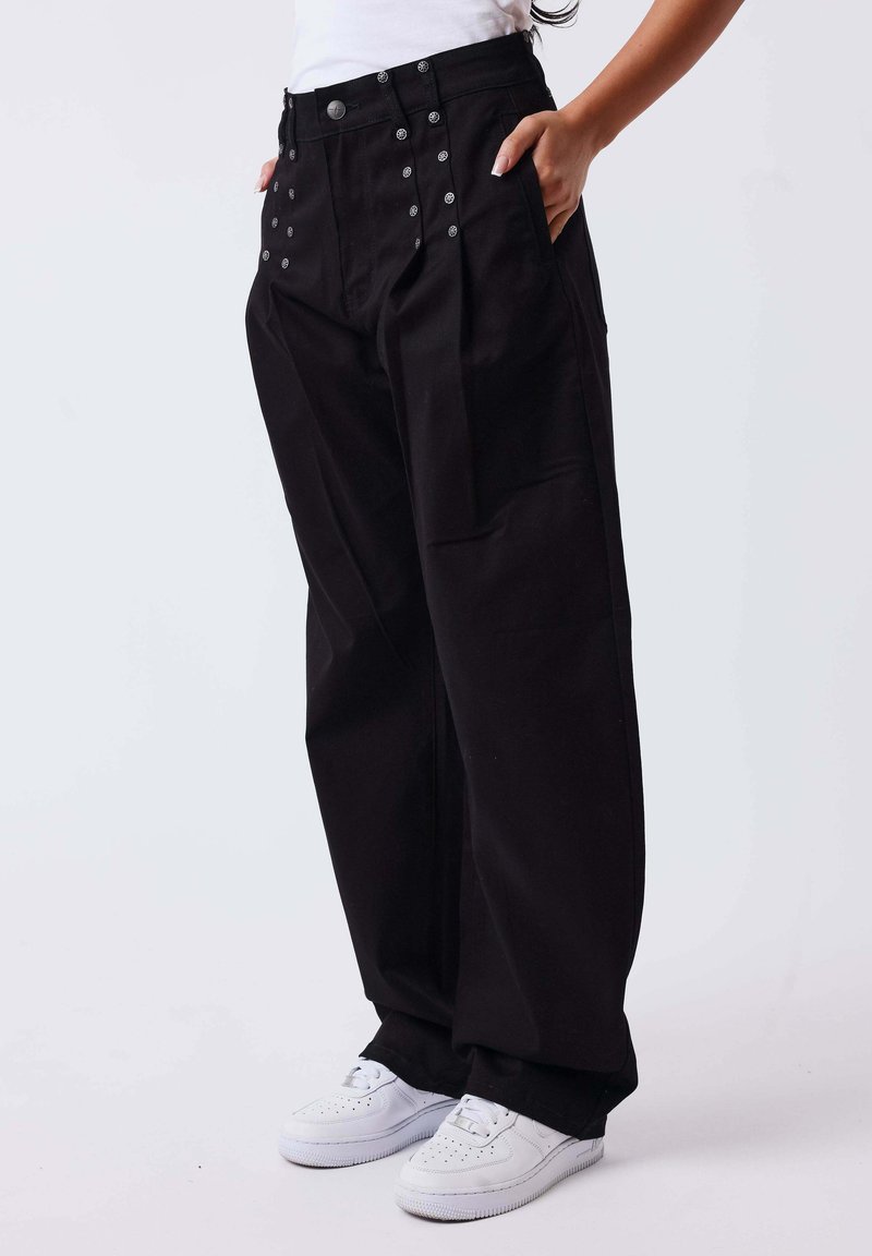 Personne portant un pantalon noir ample avec des détails de boutons et des baskets blanches, les mains dans les poches, debout devant un fond uni.