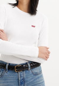 Valkoinen pitkähihaisia paita, jossa on punainen Levi's-logo rinnassa, yhdistettynä korkeavyötäröisiin sinisiin farkkuihin ja punottuun mustaan nahkavyöhön.