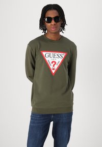 Grønn langermet genser med et hvitt og rødt trekantlogo med teksten "GUESS", kombinert med mørkeblå jeans og svarte solbriller.