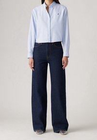 Chemise bleu clair à boutons avec manches longues et un petit logo, associée à un jean bleu foncé taille haute à jambes larges et des chaussures argentées.