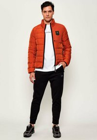Giacca arancione imbottita con dettagli neri, zip completa, colletto alto, indossata sopra una maglietta bianca con logo. Pantaloni neri a sigaretta e sneaker.