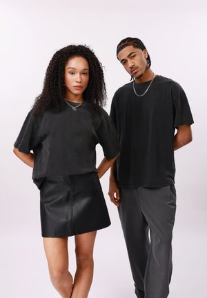 Dva mlada odrasla sta sta za preprostim ozadjem, oblečena v črne prevelike majice; ženska nosi črno krilo, moški pa sive hlače.