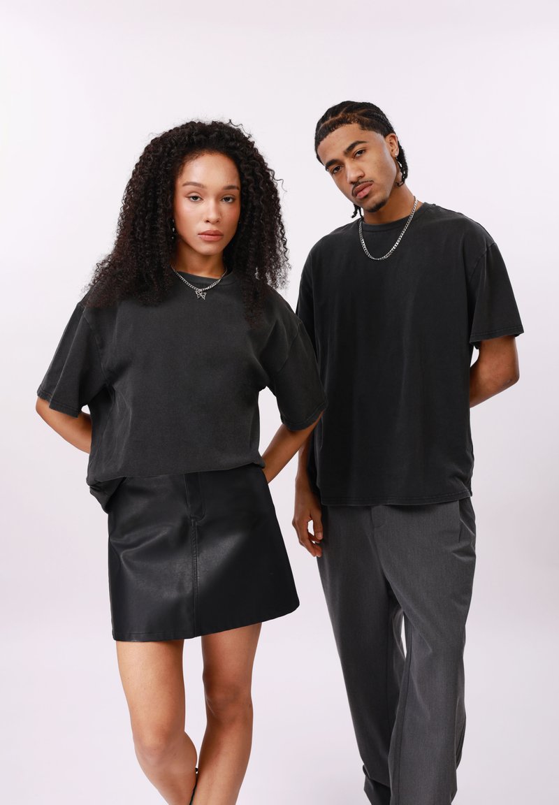 Δύο νέοι ενήλικες ποζάρουν μπροστά σε απλό φόντο, φορώντας μαύρα φαρδιά μπλουζάκια. Η γυναίκα φοράει μαύρη φούστα, ο άνδρας γκρι παντελόνι.