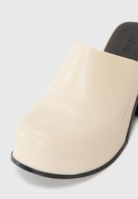 Mule de culoare crem, confecționată dintr-un material neted și lucios. Prezentă o vârful rotund, un contur negru pe baza și o talpă rezistentă.
