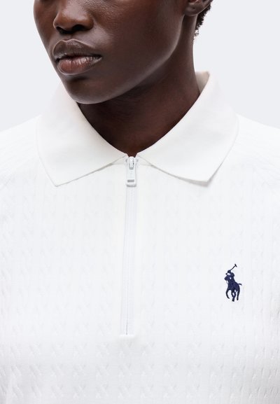 Personne portant un polo tricoté blanc à fermeture éclair, avec un col et un petit logo cheval et cavalier bleu foncé sur la poitrine.