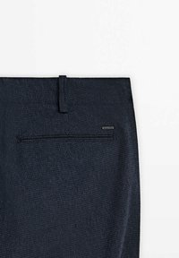 Pantalon en tissu texturé bleu foncé avec une unique poche passepoilée à l'arrière et une petite étiquette de marque au-dessus de la poche sur le côté droit.