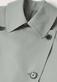 Gros plan sur un trench-coat gris clair montrant un col pointu et trois boutons noir et blanc sur la patte de boutonnage avant.