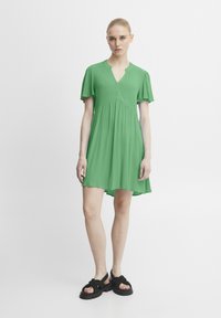 Robe courte verte avec un décolleté en V, style skater, manches bouffantes courtes, fabriquée en tissu léger, associée à des sandales à plateforme noires.