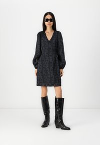 Robe noire avec des manches bouffantes et un décolleté en V, présentant un motif subtil. Associée à des bottes noires brillantes en cuir à motif crocodile, montantes jusqu'aux genoux.