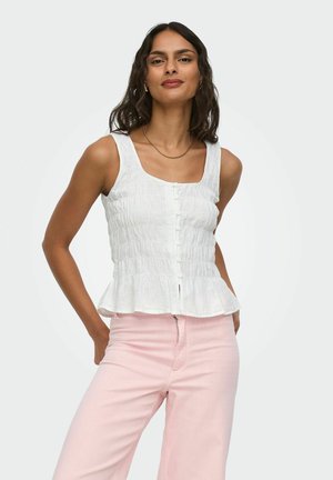 Femme aux cheveux ondulés portant de petites boucles d'oreilles créoles, un chemisier blanc sans manches à boutons et un pantalon rose clair, debout les mains derrière le dos.