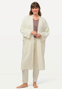 Lunga veste bianca con maniche larghe e tasche, indossata sopra una maglietta leggermente viola e pantaloni coordinati, realizzata in un tessuto morbido.
