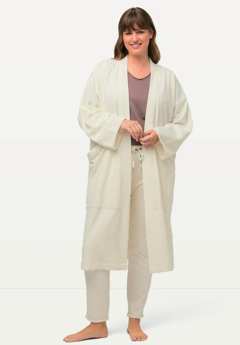 Lunga veste bianca con maniche larghe e tasche, indossata sopra una maglietta leggermente viola e pantaloni coordinati, realizzata in un tessuto morbido.