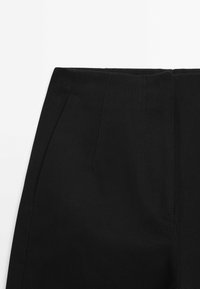 Gros plan d'un pantalon en tissu noir avec une poche latérale visible et des coutures détaillées sur fond blanc.