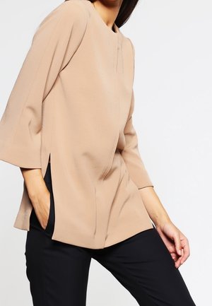 Vrouw draagt een beige, losvallend shirt met lange mouwen in combinatie met een zwarte broek, poseert met één hand in de zak tegen een witte achtergrond.