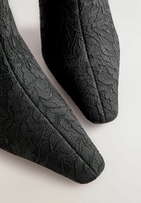 Černé krajkové kotníkové boty s špičatou špičkou. Materiál vykazuje texturovaný květinový vzor a hladký povrch.