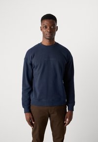 Navy blue sweatshirt with a crew neck, ribbed cuffs, and hem. Features subtle "SCOTCH & SODA" logo in the center. Worn with brown pants.