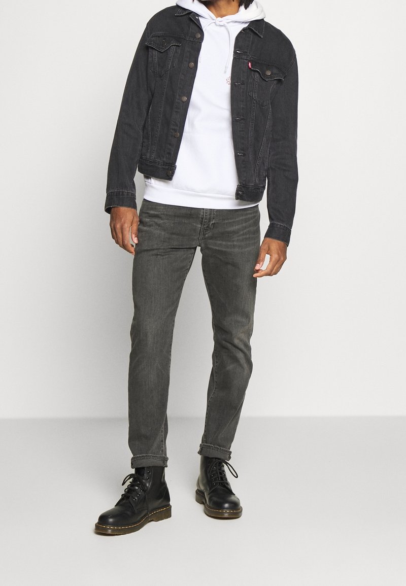 Giacca di jeans nera sopra un hoodie bianco, abbinata a jeans grigi e stivali neri. Dettagli notevoli includono cuciture e dettagli con bottoni.
