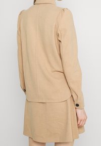 Giacca beige con maniche a sbuffo realizzata in un tessuto morbido. Presenta un colletto alto, un dettaglio sulle cuciture posteriori e un polsino con bottone. Accompagnata da un vestito abbinato.