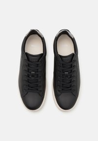 Baskets en cuir noir avec une texture lisse, bout arrondi, lacets plats, et une semelle en caoutchouc blanche ; présentent un logo subtil sur le côté et la languette.