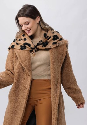 Cappotto teddy marrone e morbido con una sciarpa beige drappeggiata con macchie nere. Indossato sopra un maglione beige a coste e pantaloni marroni a vita alta.