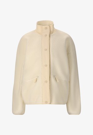 Jonge vrouw staat met handen in haar zakken, draagt een crèmekleurige fleecejack, zwarte Les Arcs fietsshorts, witte sokken en zwarte sneakers tegen een witte achtergrond.