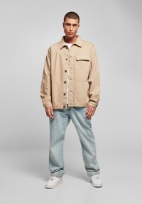Beige lerretsjakke med krage, frontlukking med knapper, en brystlomme, to frontlommer og en avslappet passform, kombinert med lyseblå jeans.