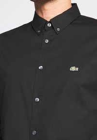 Czarna koszula na guziki z bawełnianej tkaniny, z klasycznym kołnierzem i małym, zielonym haftowanym logo krokodyla. Guziki są szare.