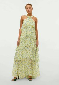 Maxi suknelė su geltonais gėlių raštais, sluoksniuota konstrukcija, mažiausiai kaklo juosta ir permatoma medžiaga. Apima plazdančią siluetą ir daugiasluoksnes raukšles.