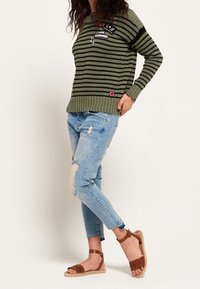 Maglione a righe verde oliva con texture a costine, abbinato a jeans chiari strappati e sandali marroni con cinturini e dettagli intrecciati.