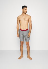 Graue Sportshorts mit einem roten "UNDER ARMOUR"-Bund, ausgestattet mit einer konturierten Beuteltasche und einem elastischen, strukturierten Stofffinish.