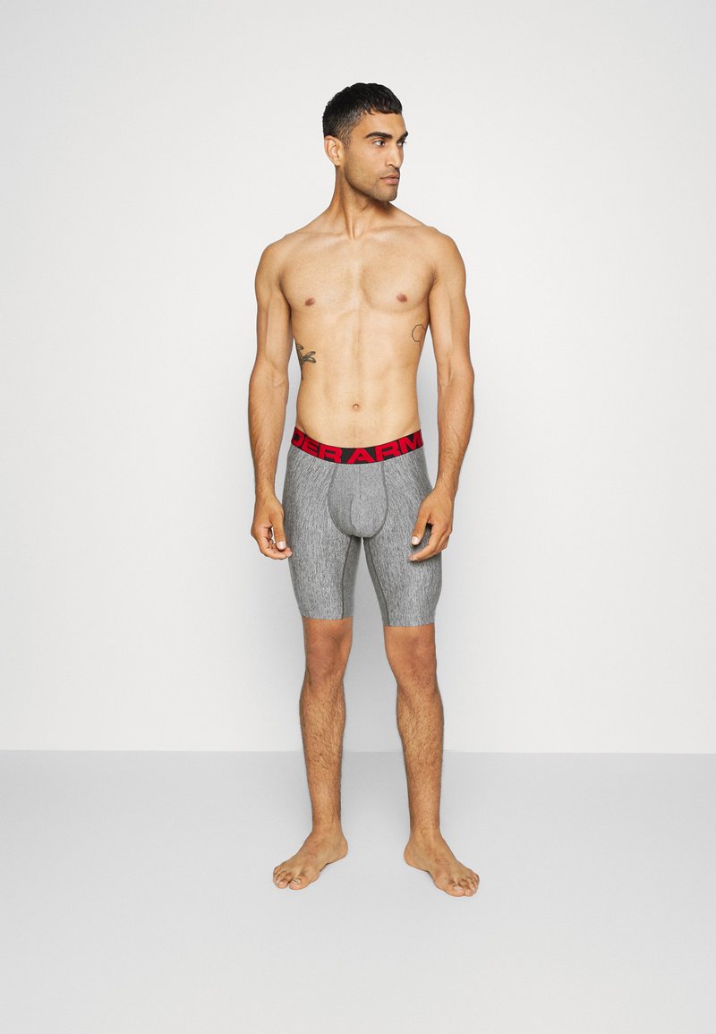 Graue Sportshorts mit einem roten "UNDER ARMOUR"-Bund, ausgestattet mit einer konturierten Beuteltasche und einem elastischen, strukturierten Stofffinish.