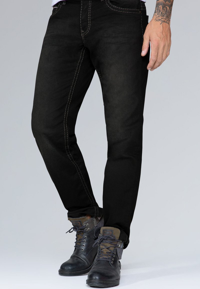 Jeans negros ajustados con costuras claras en contraste. Llevados con botas oscuras con cordones que presentan un diseño robusto y acentos texturizados.