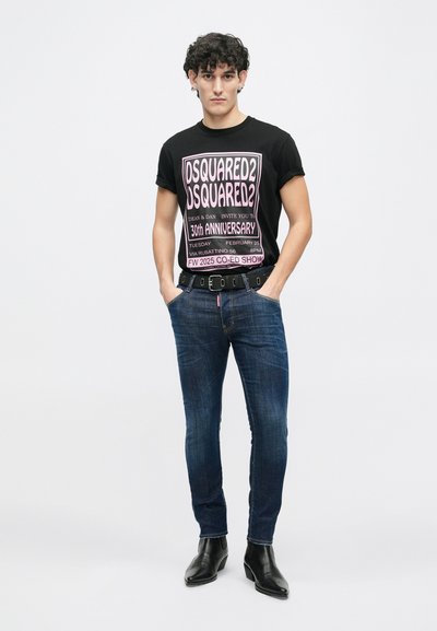 Camiseta gráfica negra con texto rosa, vaqueros de mezclilla azul y botas de tobillo de cuero negro con acabado texturizado. Cinturón con características distintivas.