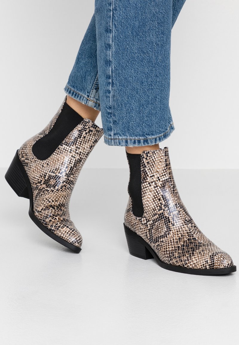 Une personne portant un jean bleu et des bottes à motif serpent beige avec des côtés en élasthanne noir et des talons empilés, se tenant sur un sol clair.