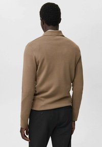 Man wearing a long-sleeve, camel-brown sweater and black pants, standing facing away against a plain light background.