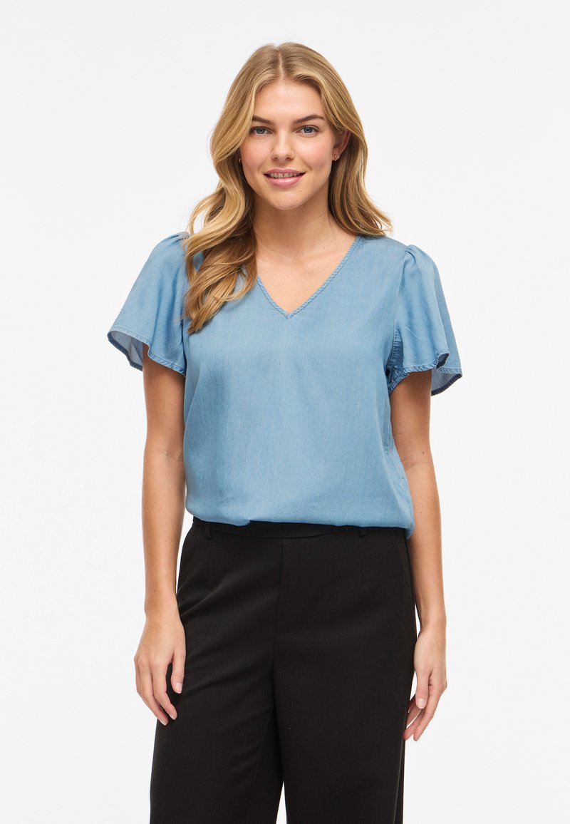 Γυναίκα με ξανθά μαλλιά, που φοράει ανοιχτό μπλε μπλούζα με κοντά μανίκια και μαύρο παντελόνι, στέκεται μπροστά σε λευκό φόντο, χαμογελώντας ελαφρώς.