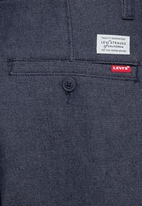 Tissu en denim bleu foncé avec une poche visible, un bouton noir et une étiquette portant la mention "Levi's" avec un accent rouge sur le bord.