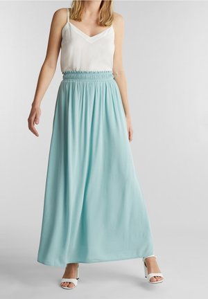 Kobieta ubrana w białą bluzkę bez rękawów i przewiewną, jasnoniebieską suknię maxi oraz białe sandały na wysokim obcasie, stojąca na jednolitym tle.