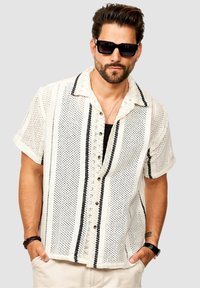 Mann mit Bart, der schwarze Sonnenbrillen trägt, ein weißes kurzärmliges Hemd mit schwarzen senkrechten Streifen und beigen Hosen anhat, mit den Händen in den Taschen.