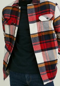 Rød, blå og gul ternet flannel-skjorte med to brystlomme, knaplukninger og et blødt, tekstureret stof. Båret over en sort top.