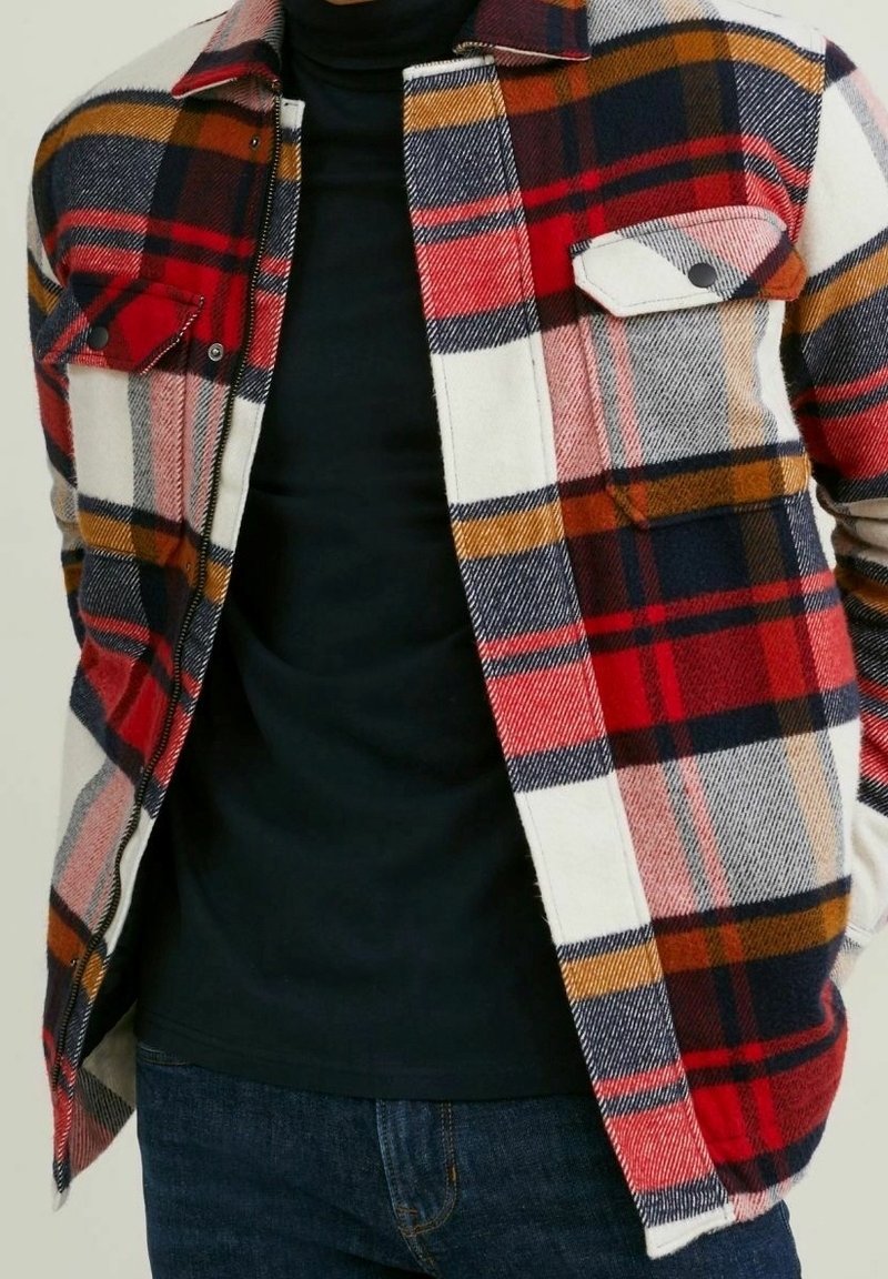 Rød, blå og gul ternet flannel-skjorte med to brystlomme, knaplukninger og et blødt, tekstureret stof. Båret over en sort top.